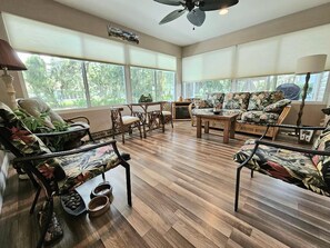 Una habitación amplia con ventanas grandes, suelo de madera y diversas opciones de asientos, incluyendo un sofá, sillas y una mesa de centro.