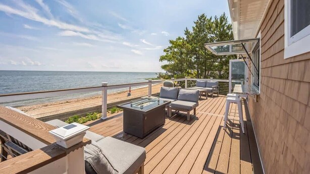 En træterrasse med udendørs møbler, et bord med glasplade og udsigt til stranden og havet.