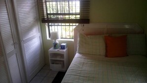 A bedroom with a bed, a nightstand, a window with blinds, and a view of greenery outside. - Henrietta's Seaview Studio (Gros Islet)