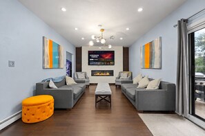 Un salon moderne avec des canapés gris, un pouf central, une cheminée et un grand écran de télévision diffusant un film.