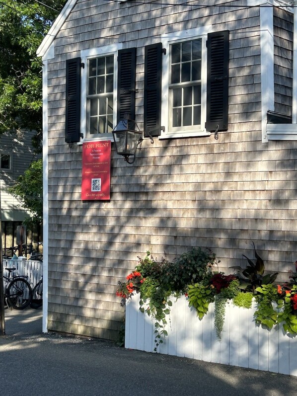 A small building with a 'For Rent' sign, a lantern, and a planter with flowers.