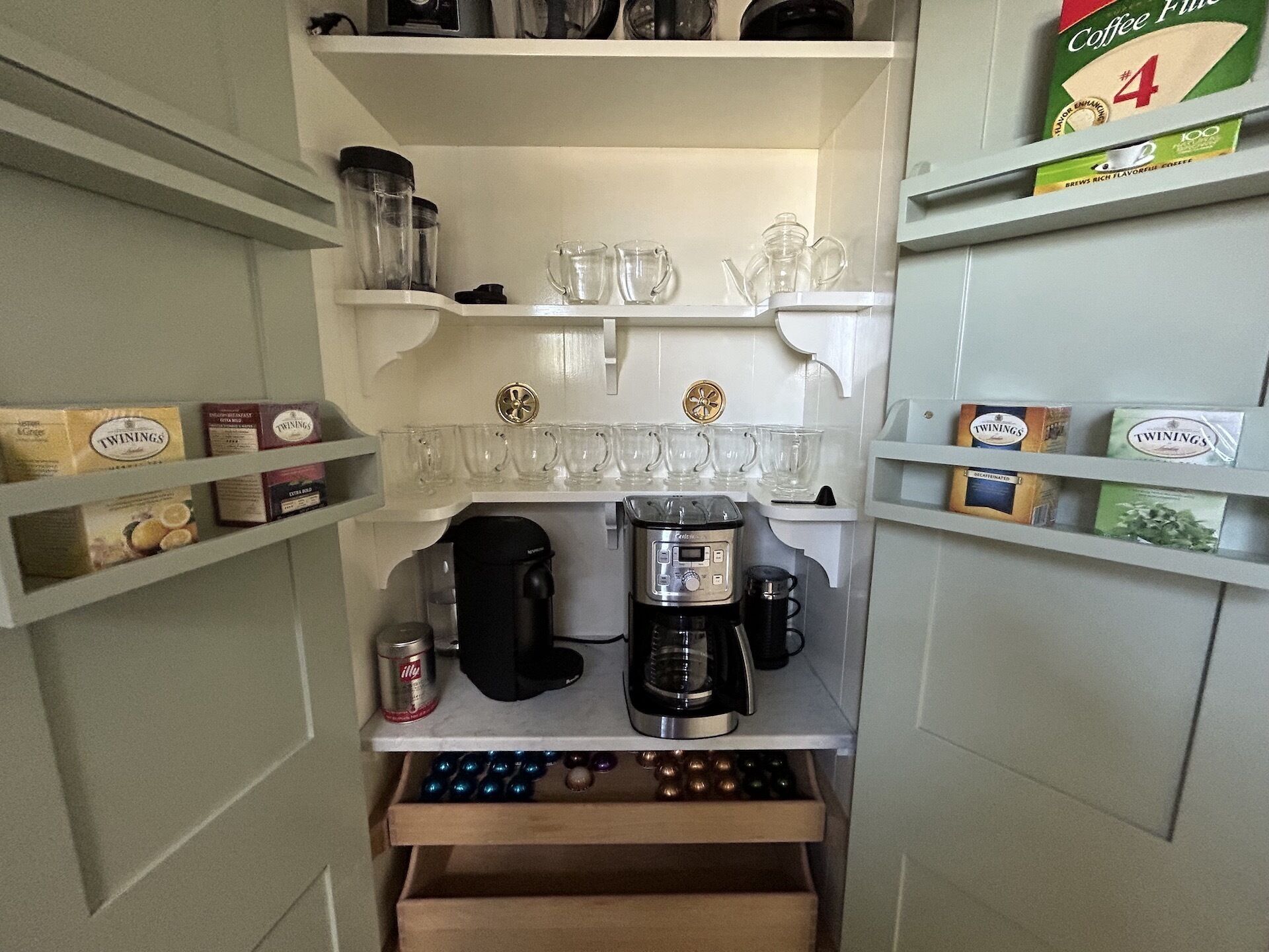 A compact kitchenette with a coffee maker, tea kettle, and various coffee pods on shelves.