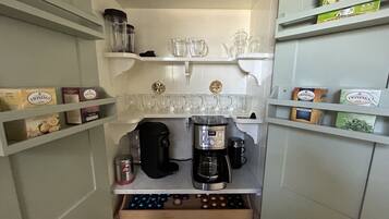 A compact kitchenette with a coffee maker, tea kettle, and various coffee pods on shelves.