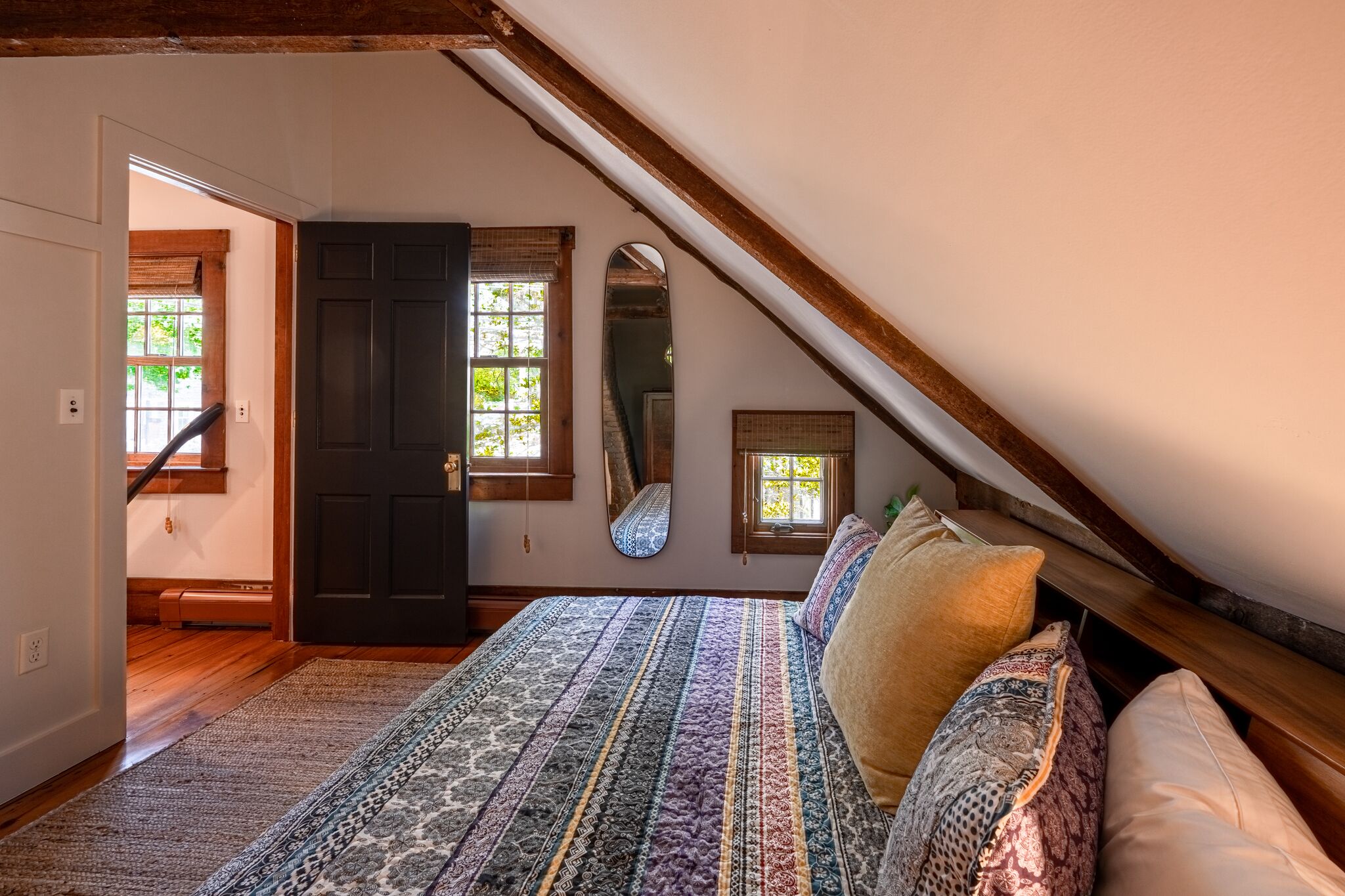Ein Schlafzimmer mit schrägen Wänden, einem Bett mit gemusterten Textilien, einem Fenster mit Blick und einer Tür zu einem weiteren Raum.