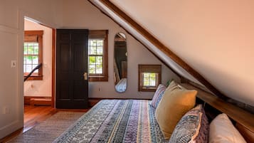 Ein Schlafzimmer mit schrägen Wänden, einem Bett mit gemusterten Textilien, einem Fenster mit Blick und einer Tür zu einem weiteren Raum.
