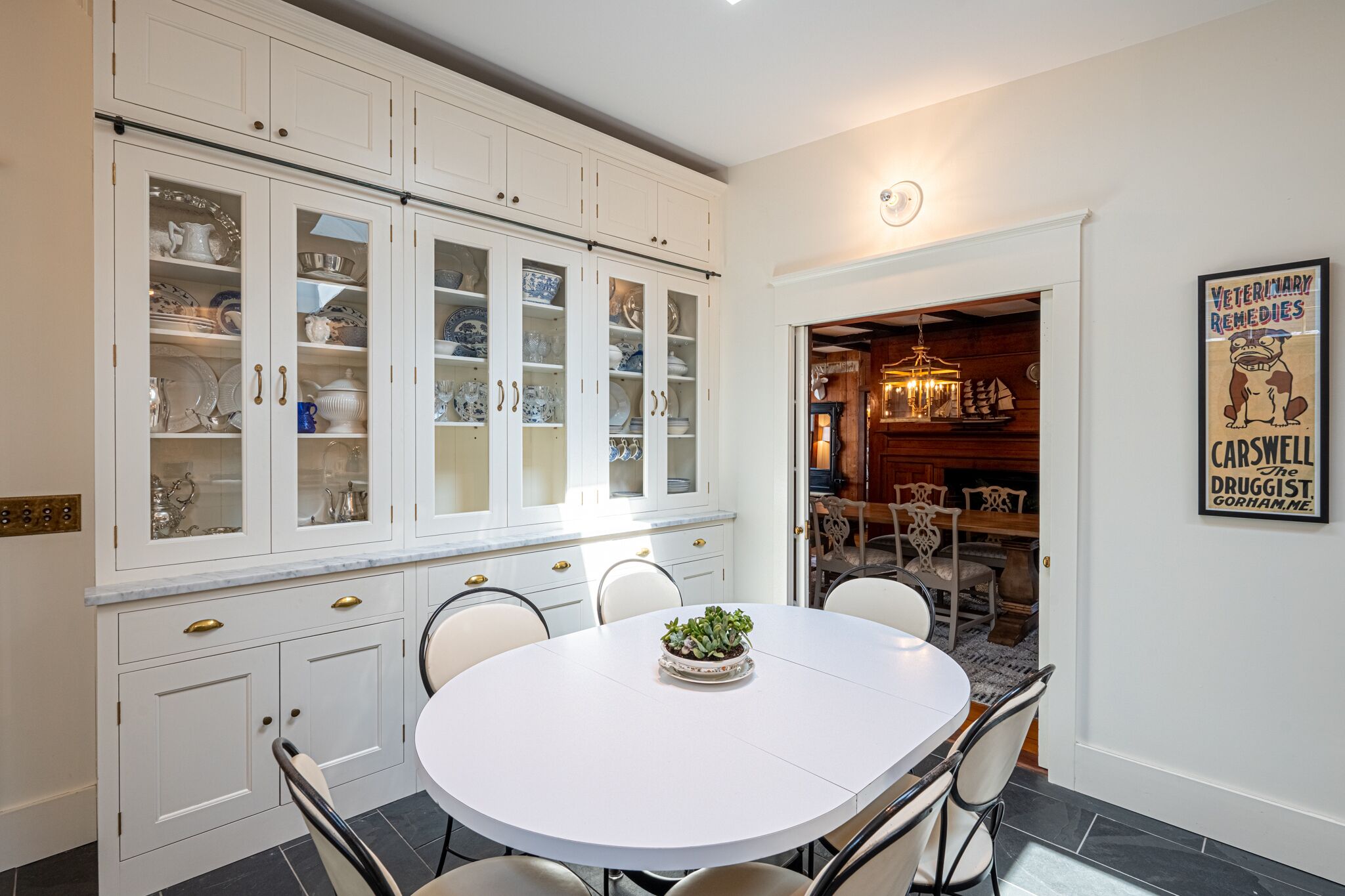 A dining area with a white table and chairs, a potted plant, and a cabinet with glass doors containing dishes.