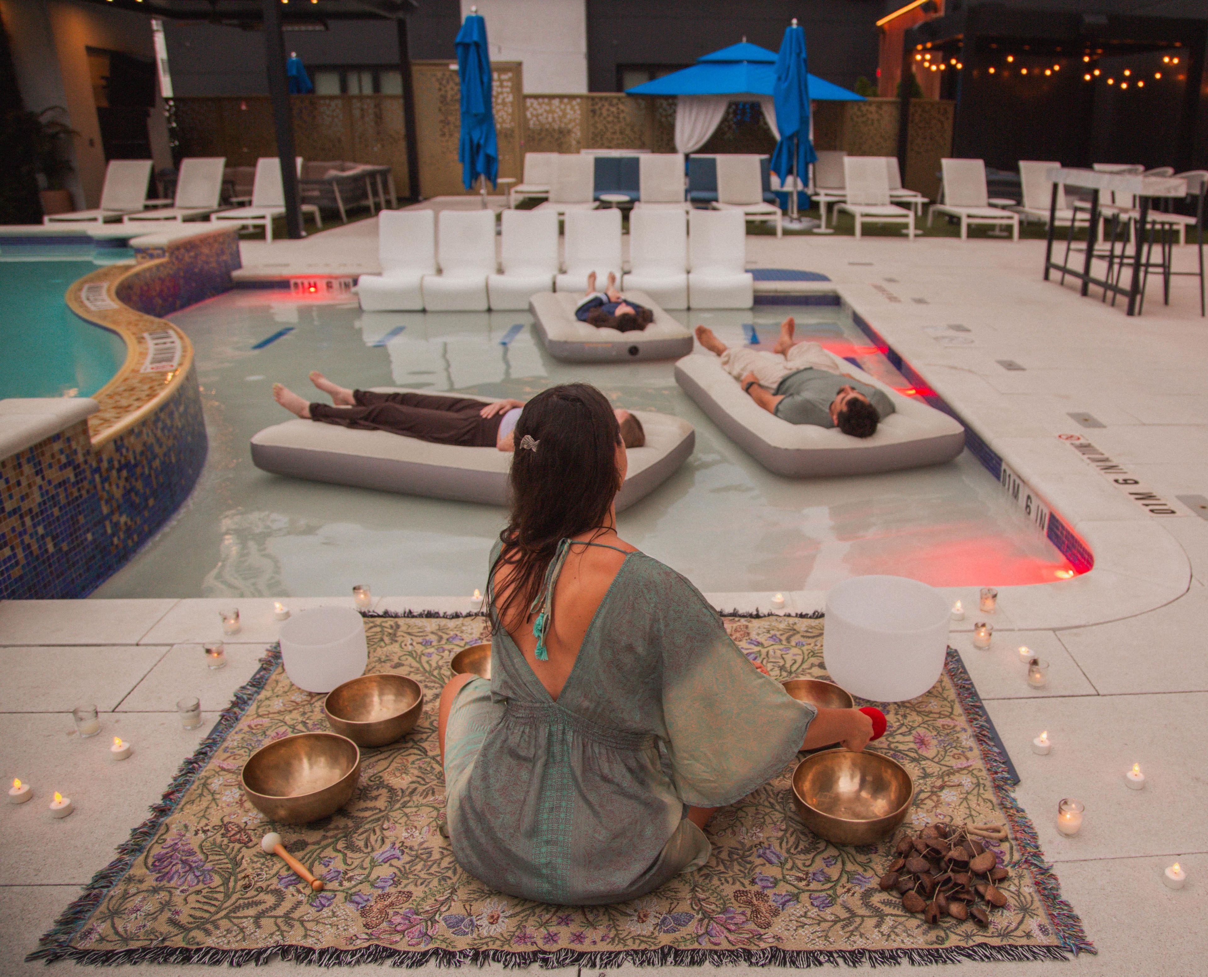 Un área de relajación junto a la piscina con sillones de descanso y una persona meditando con cuencos.
