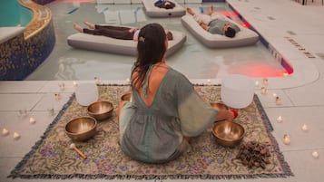 Un área de relajación junto a la piscina con sillones de descanso y una persona meditando con cuencos.