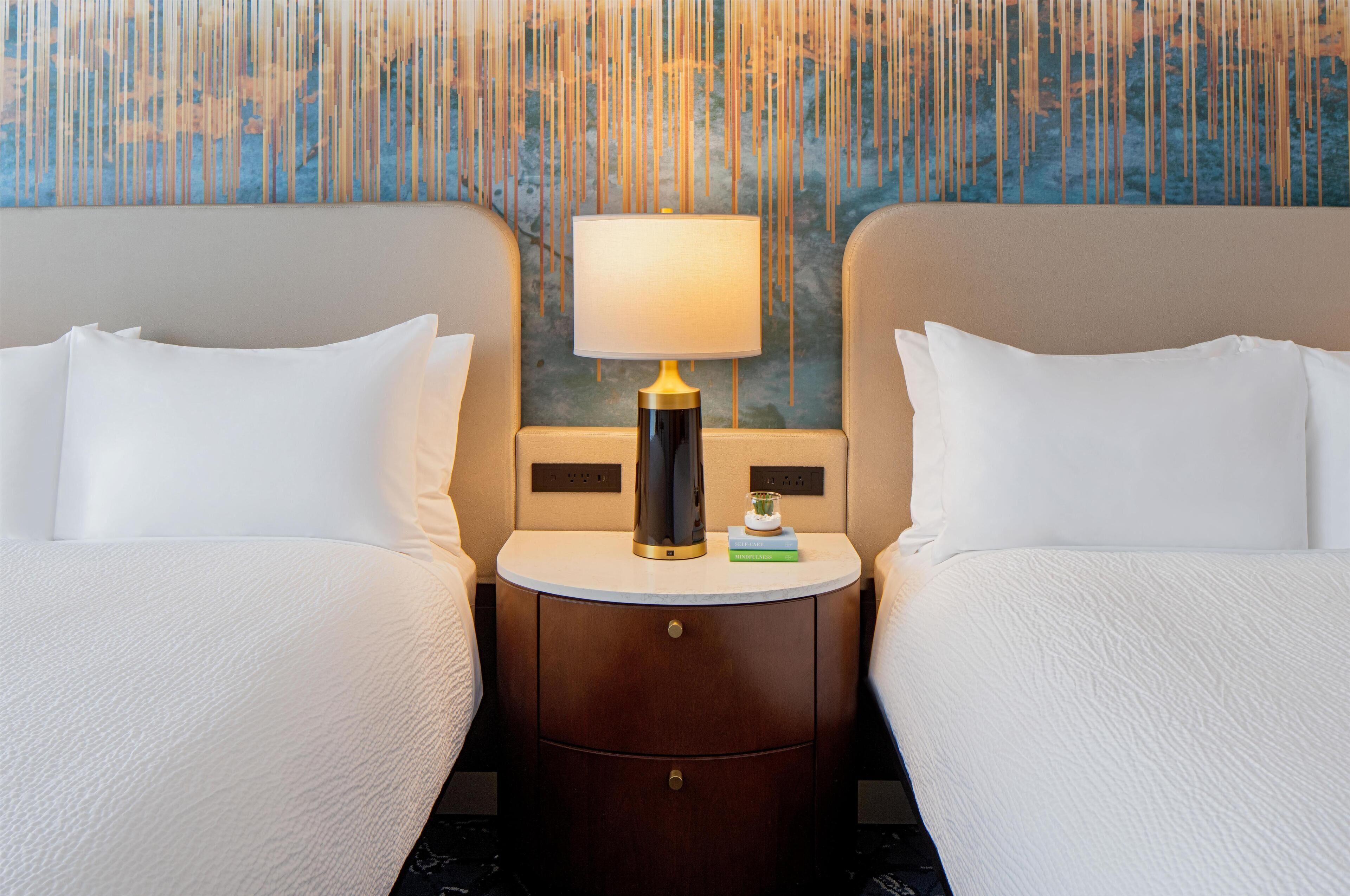 A hotel room with two beds, a bedside table with a lamp, and a decorative wall.
