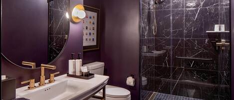 A bathroom with a white sink, gold faucets, a white toilet, and a glass-enclosed shower with a marble wall.