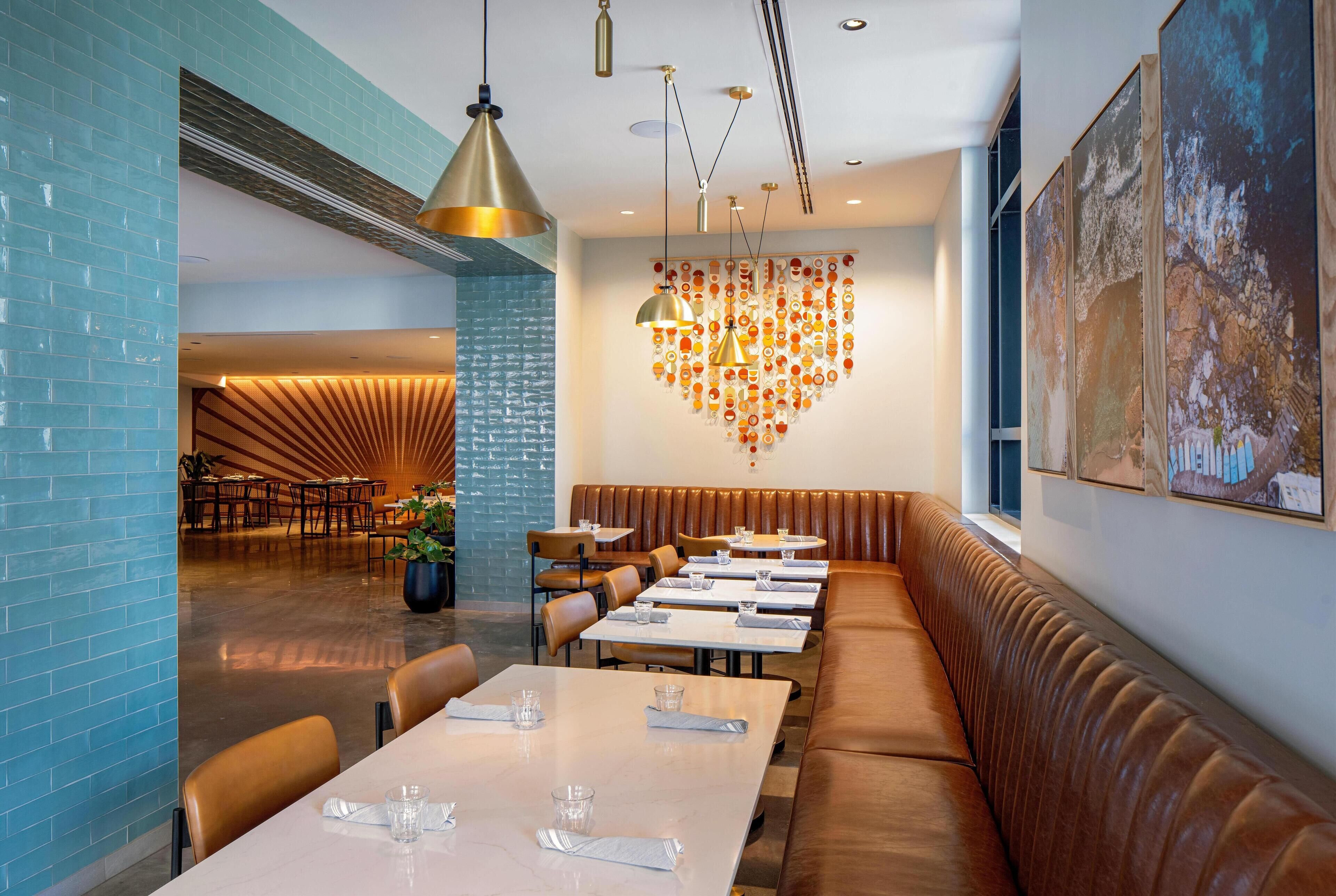 A modern restaurant interior with a long brown leather booth, multiple tables, and hanging pendant lights.