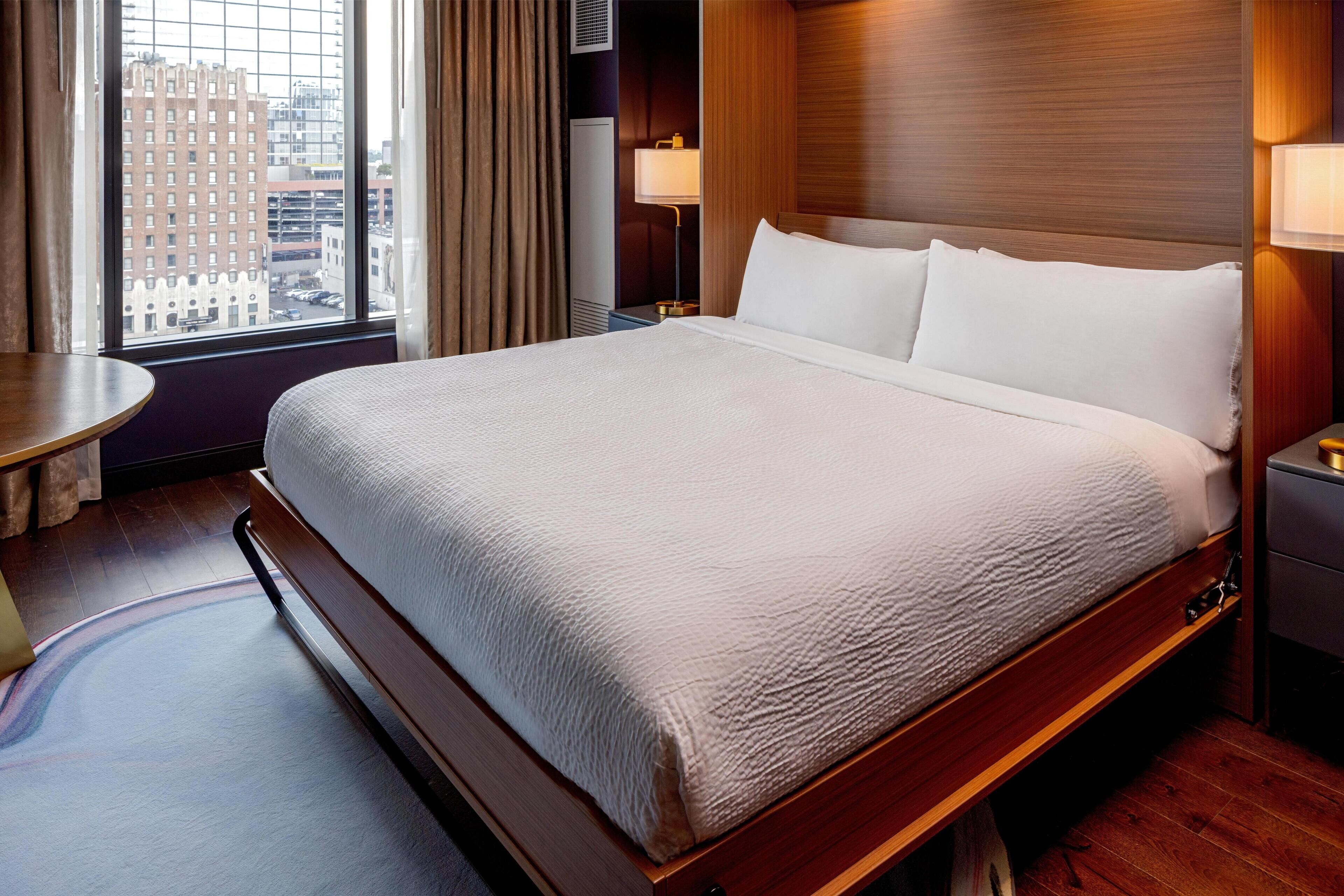 Habitación de hotel con una cama grande, una mesita de noche, una lámpara y vista a la ciudad desde la ventana.