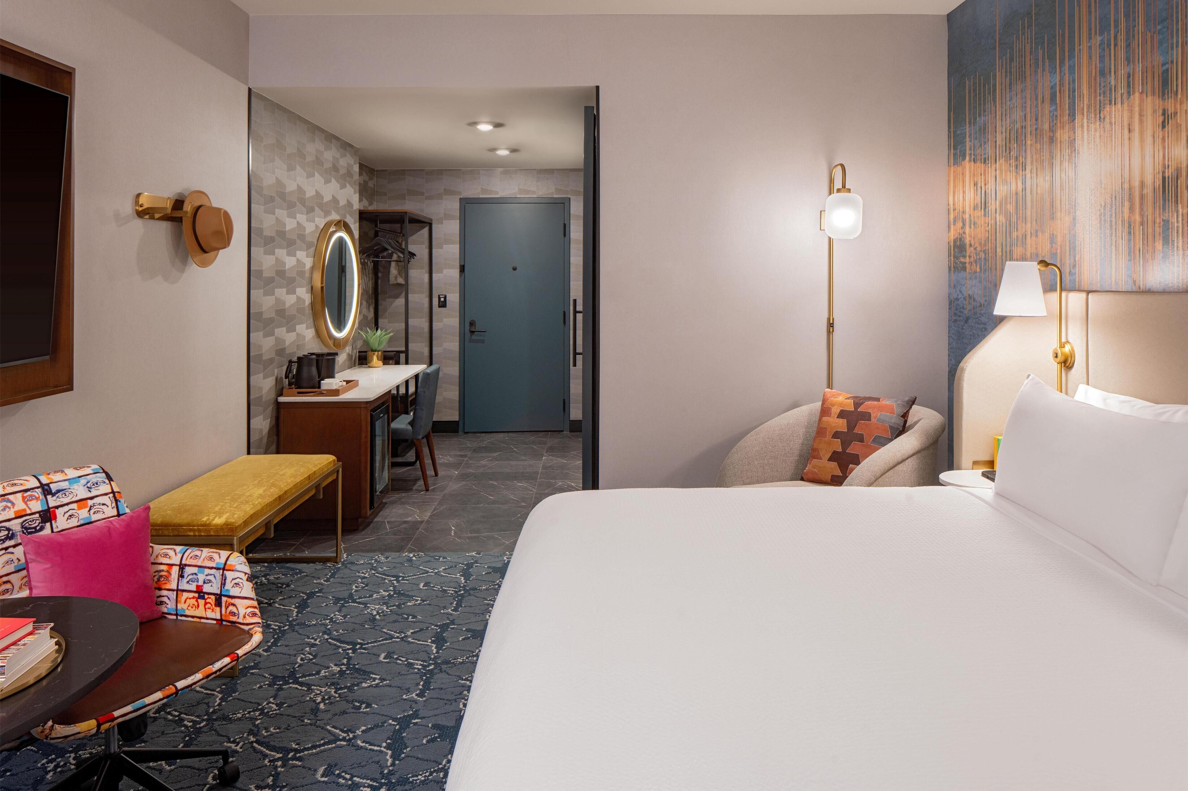 A hotel room with a large bed, a desk with a chair, a small table with a chair, a bench, a mirror, and a door leading to a bathroom.