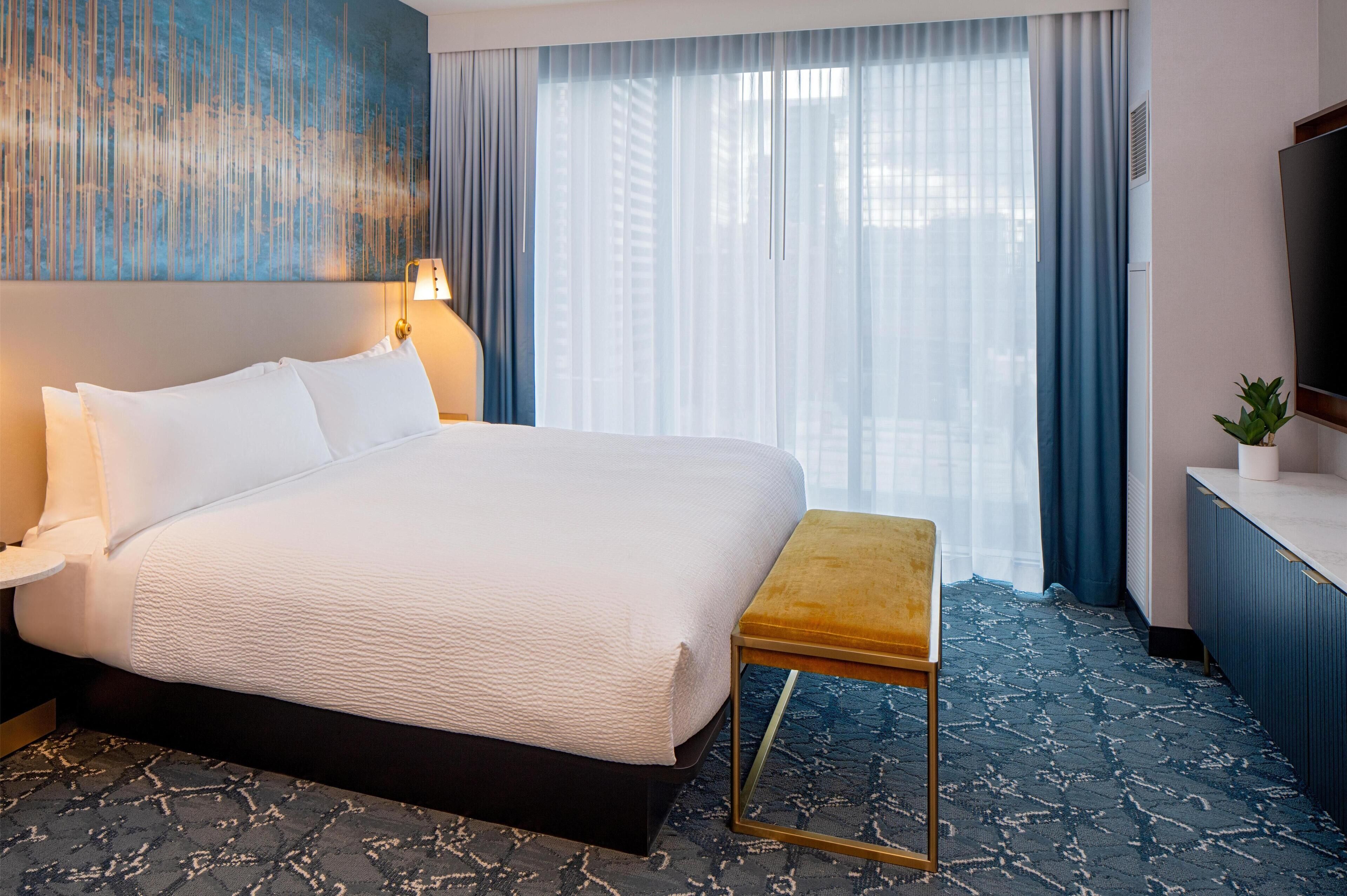 A hotel room with a large bed, a small table, a TV, and a window with curtains.