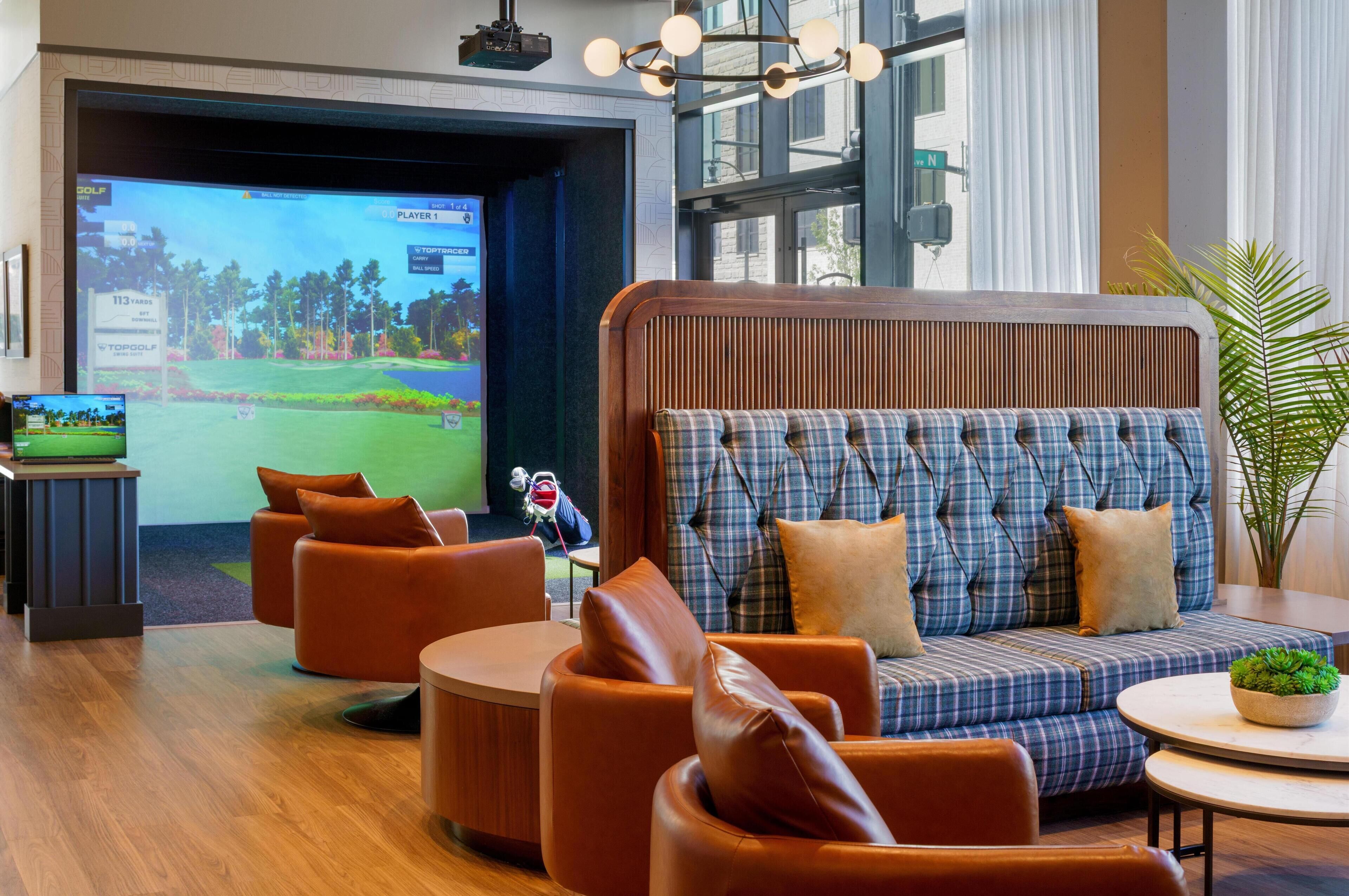 Ein moderner Lounge-Bereich mit einem großen Bildschirm, der ein Golfspiel zeigt, bequemen Sitzgelegenheiten und Blick ins Freie.