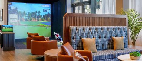 A modern lounge area with a large screen displaying a golf game, comfortable seating, and a view of the outdoors.