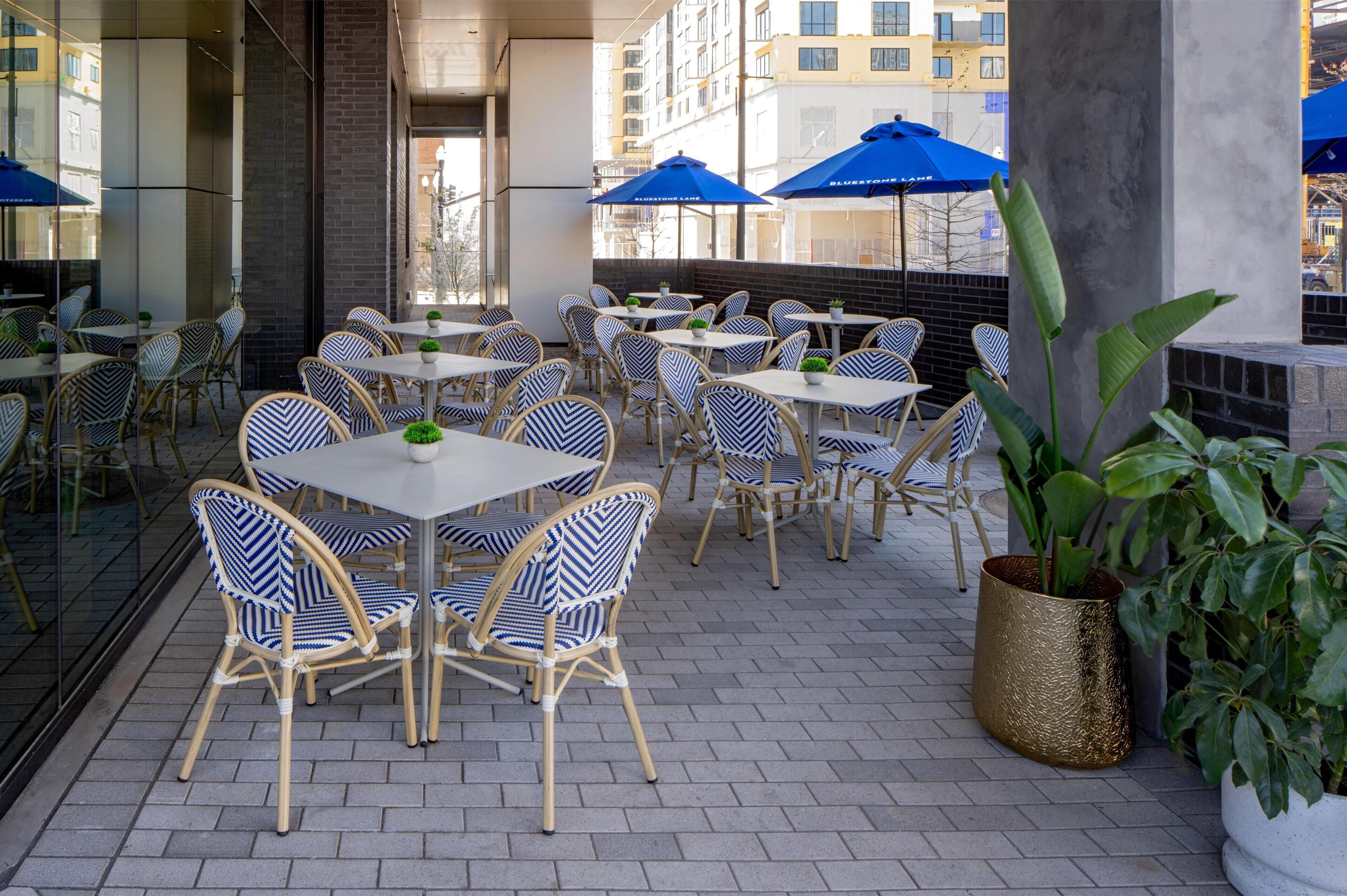 Un área de café al aire libre con sillas y mesas de mimbre, sombrillas azules y plantas en macetas.