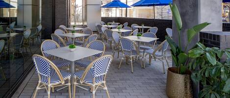 Un área de café al aire libre con sillas y mesas de mimbre, sombrillas azules y plantas en macetas.