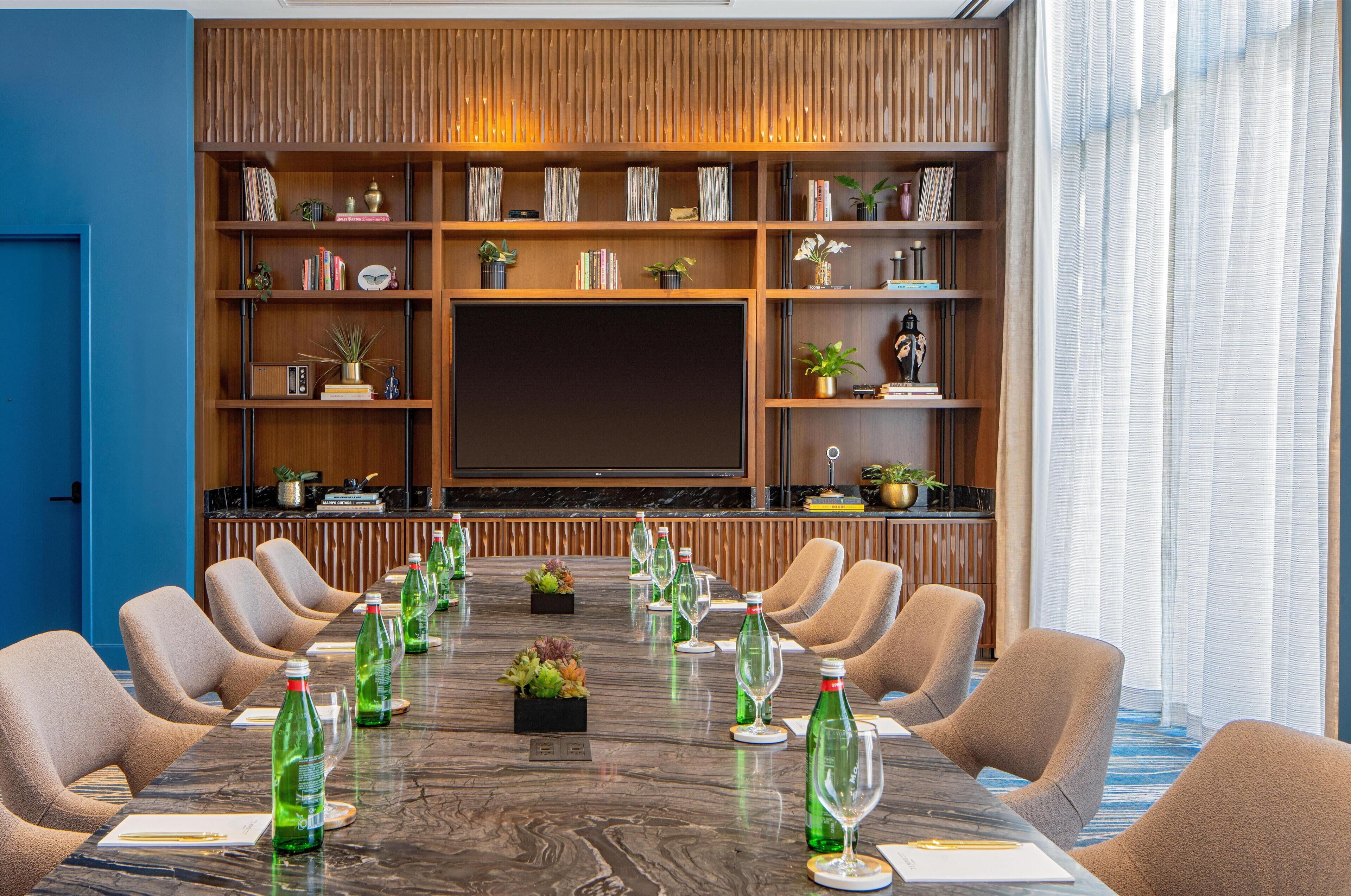 A conference room with a long table, chairs, a television screen, and a bookshelf with various items.