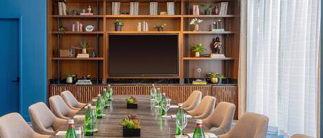 A conference room with a long table, chairs, a television screen, and a bookshelf with various items.