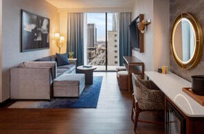 A modern hotel room with a sofa, a small coffee table, a desk with a chair, a large mirror, and a view of the city through the window. - Tempo By Hilton Nashville Downtown (Nashville)