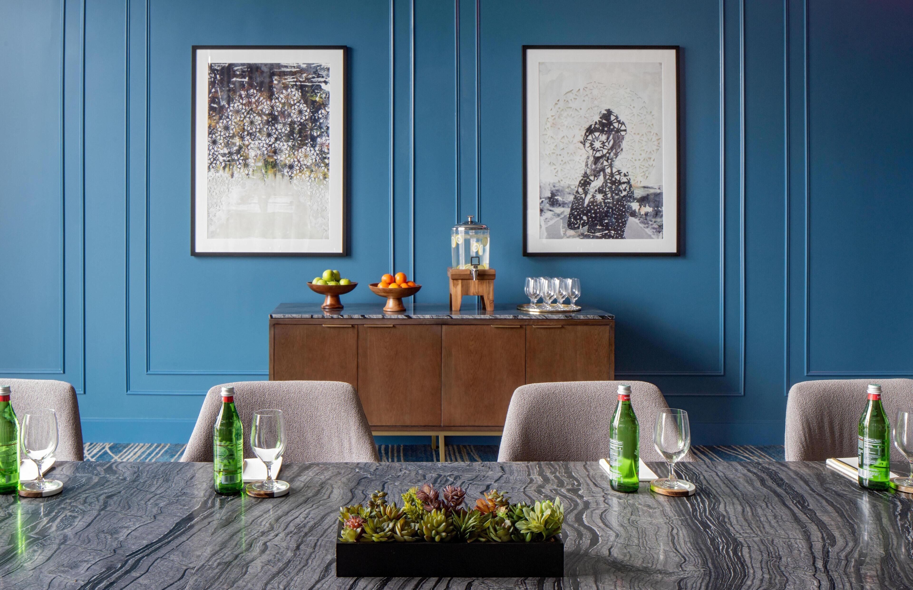 A dining area with a blue wall, two framed artworks, a wooden cabinet, and a table with chairs and green bottles.