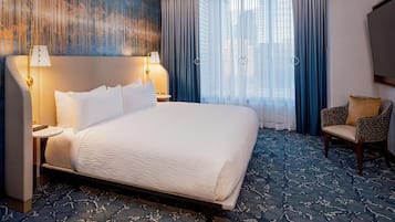 Una habitación de hotel con una cama grande, dos lámparas de noche, una silla, un ventanal con cortinas y una pared con líneas verticales.
