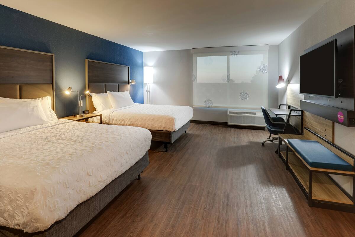 Ein Hotelzimmer mit zwei Betten, einem Schreibtisch, einem Stuhl, einem Fernseher und einem großen Fenster.