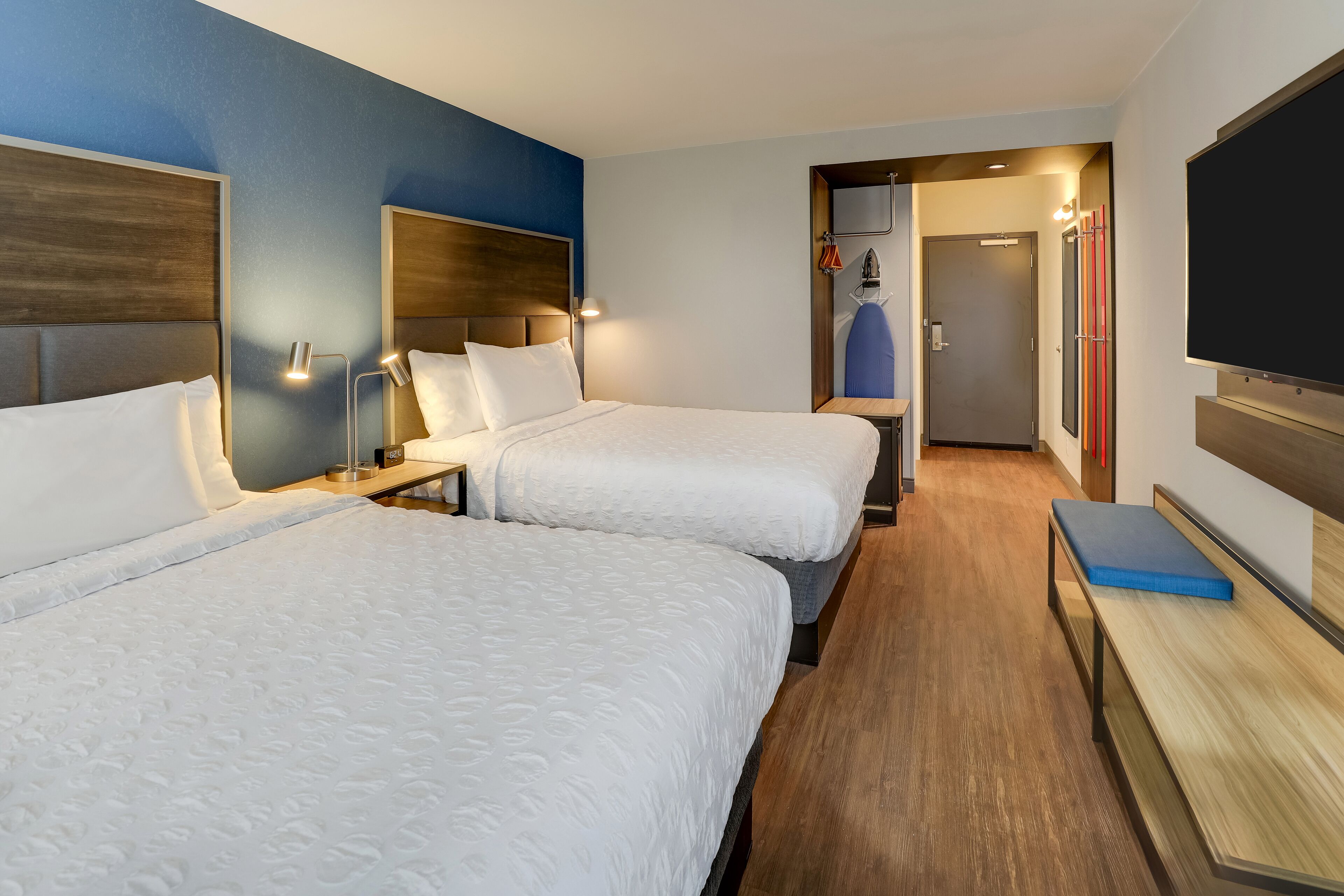 A hotel room with two beds, a TV, a desk, and a bench.