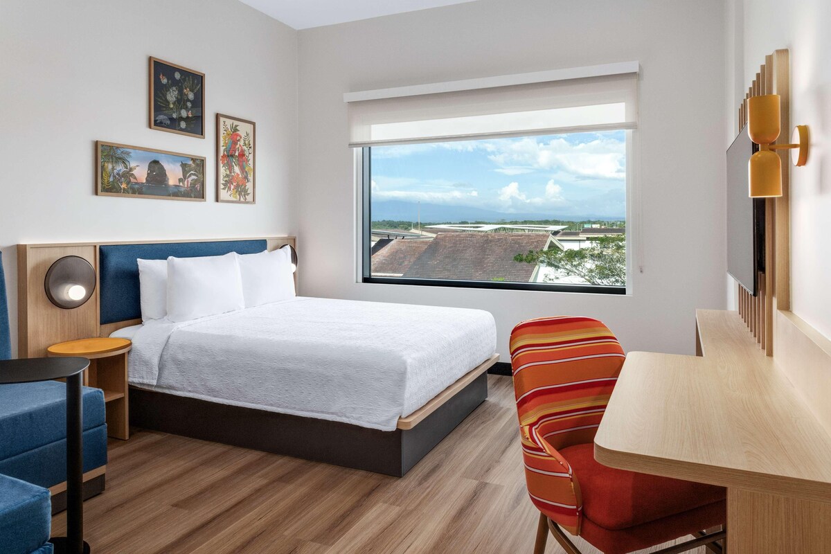 Une chambre d’hôtel moderne dotée d’un grand lit, d’un bureau, d’une chaise et d’une grande fenêtre donnant sur la ville.