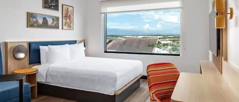 Habitación de hotel con cama, escritorio, silla, ventana grande con vista a la ciudad y decoración en la pared.