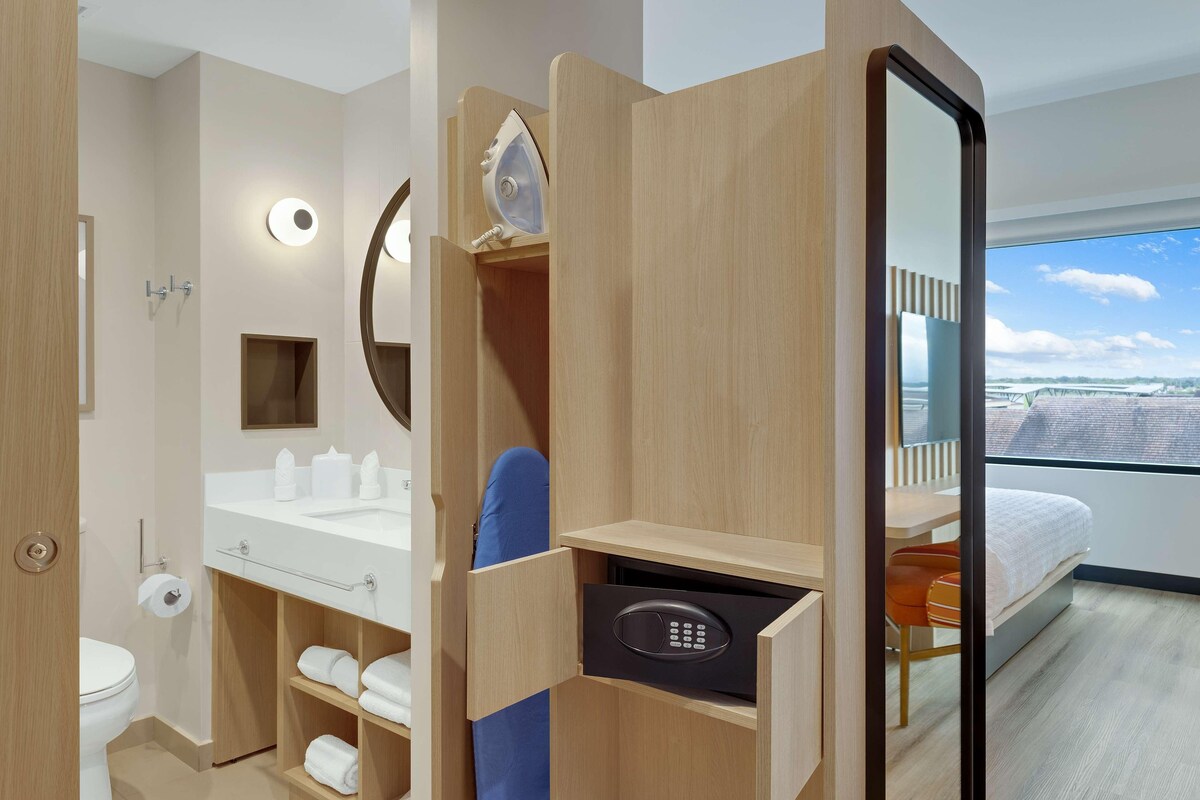 Une chambre d’hôtel moderne dotée d’une coiffeuse en bois, d’une douche intégrée, d’un grand miroir, d’un lit avec vue et d’un panneau de commande.
