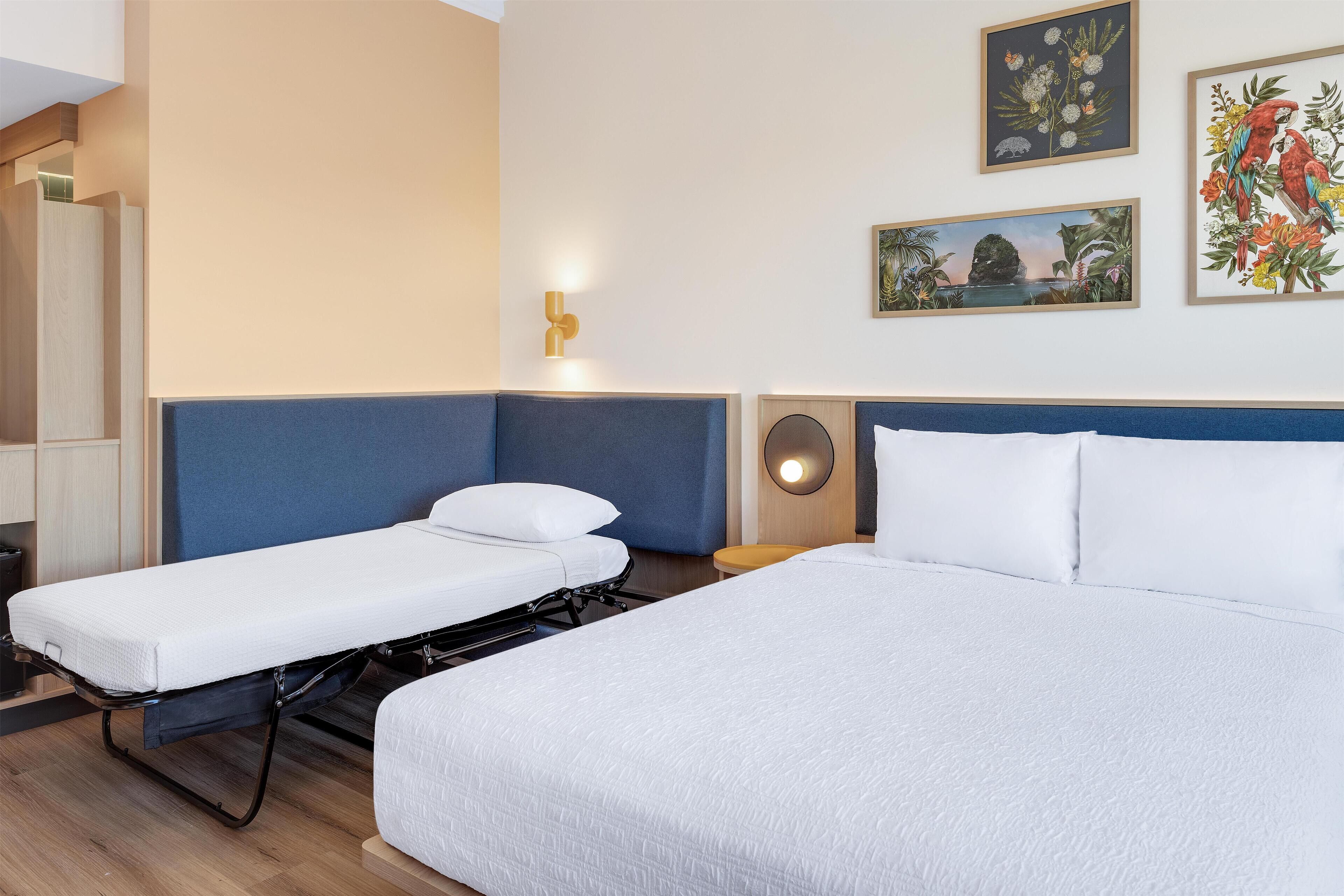 Une chambre d’hôtel avec un grand lit, un lit plus petit et un bureau avec une chaise. Des appliques murales et des œuvres d’art encadrées ornent les murs.