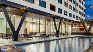 A swimming pool with lounge chairs and a bar area, surrounded by a modern building with large windows and a covered seating area.