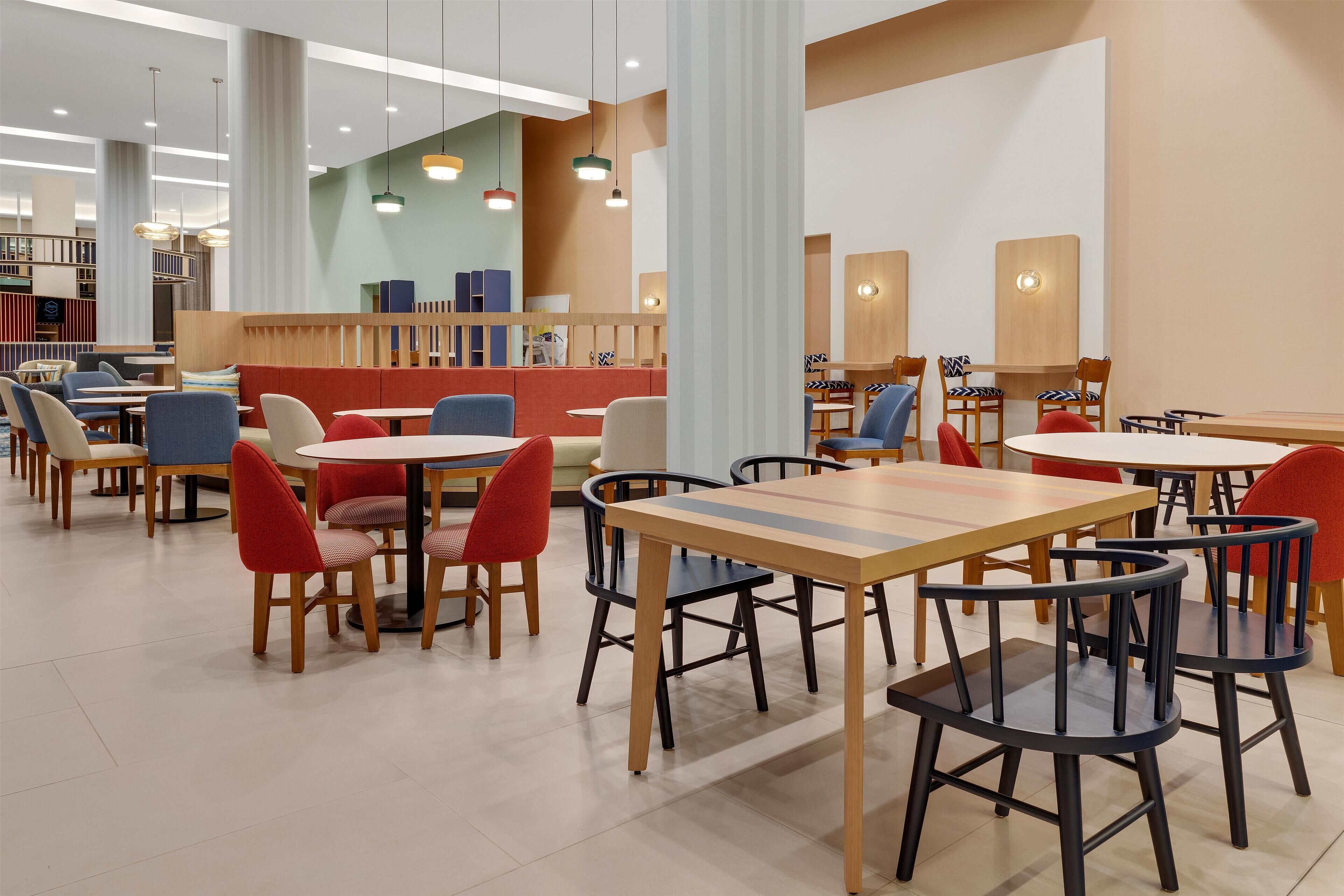 Un comedor moderno con varias mesas y sillas, un área de estar central con un banco rojo e iluminación colgante.