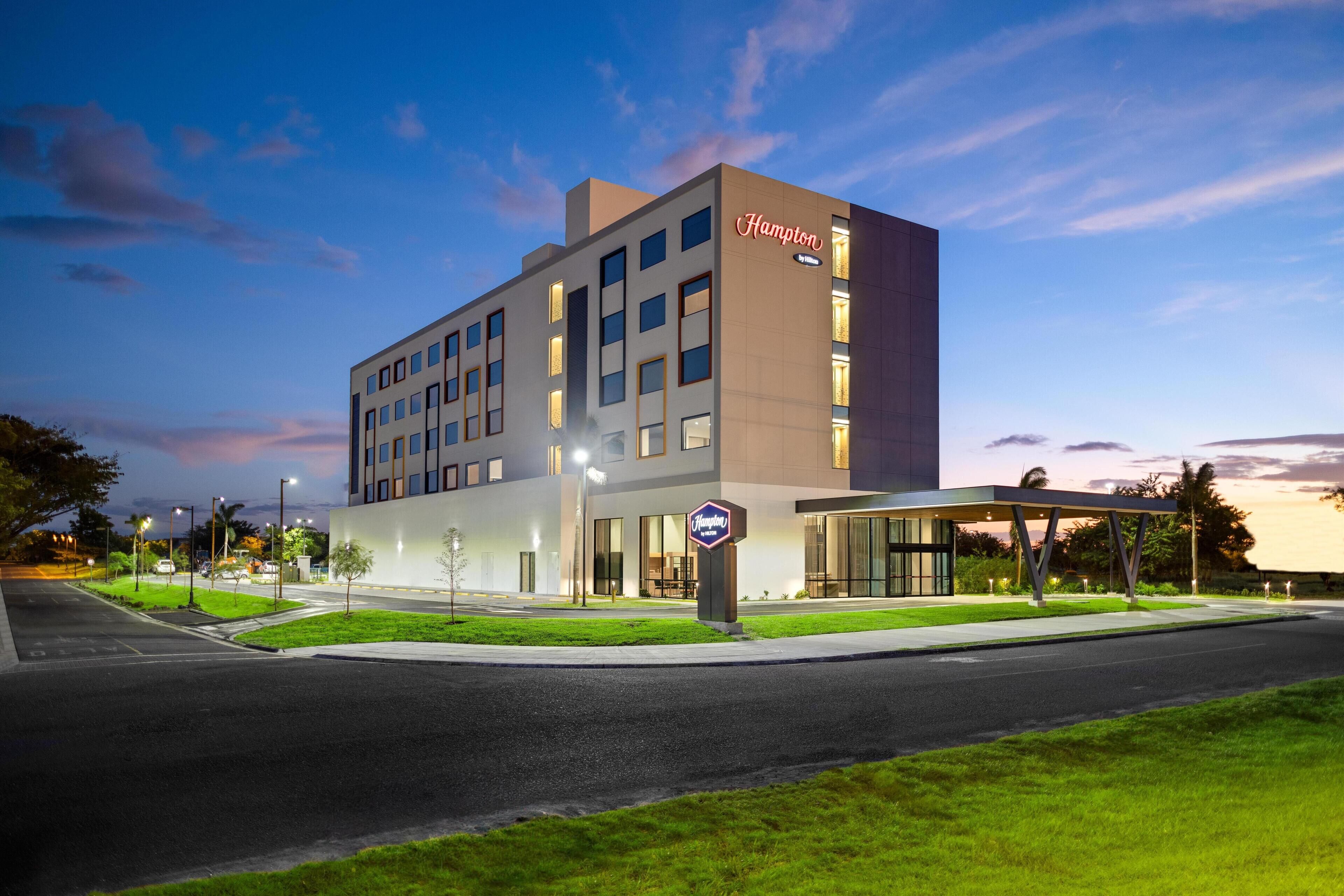 A modern hotel with a prominent 'Hampton' sign, featuring a clean, rectangular design with multiple windows and a flat roof.