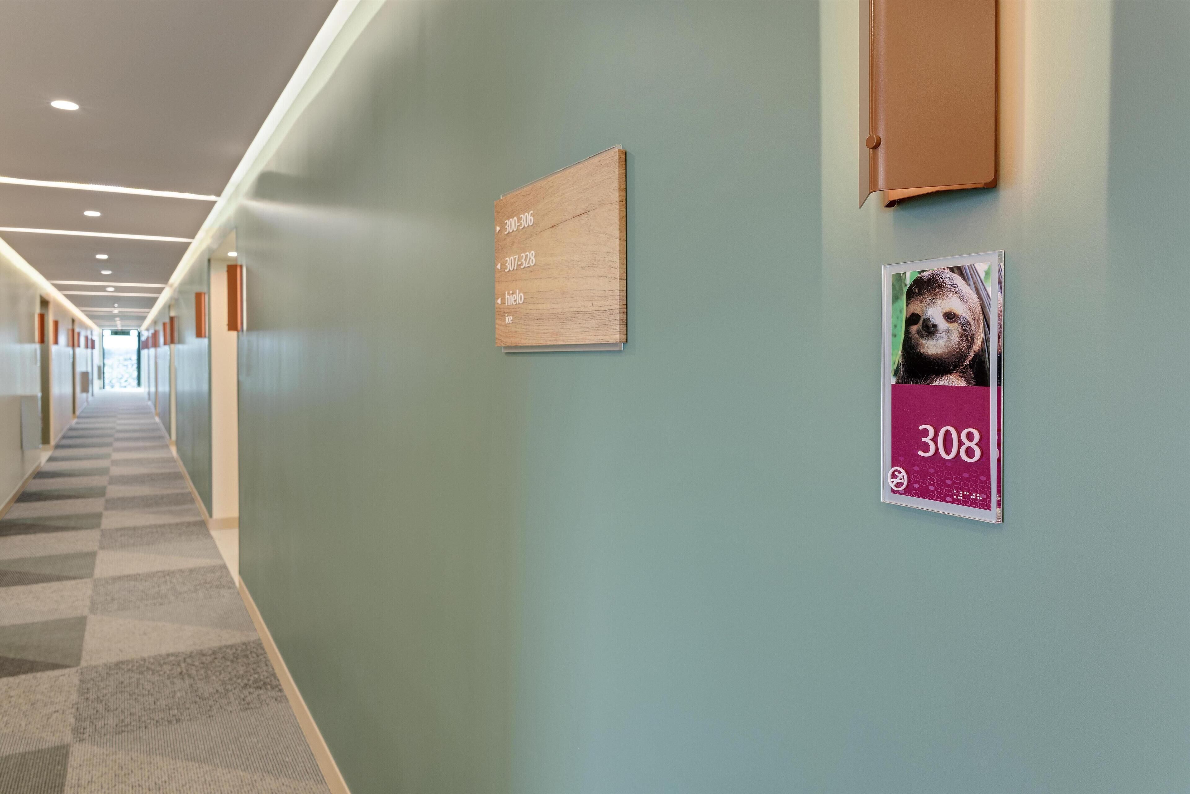A hallway with a checkered carpet, a wooden sign with room numbers, and a sloth poster.