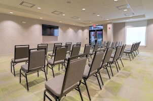 Un salón de conferencias con filas de sillas plegables, un televisor de pantalla plana montado en la pared y un gran ventanal que proporciona luz natural.