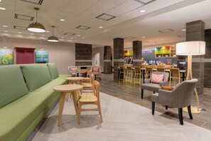 A modern hotel lobby with a green sofa, wooden chairs, a bar area, and a reception desk. - Home2 Suites By Hilton Alcoa Knoxville Airport (Alcoa)