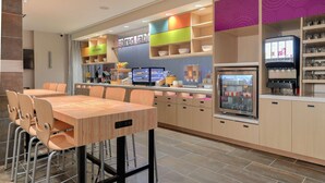 A modern restaurant with a wooden dining area, colorful shelving, and a self-service beverage station. - Home2 Suites By Hilton Alcoa Knoxville Airport (Alcoa)