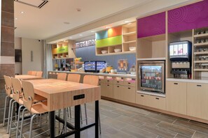 Un restaurante moderno con zona de comedor de madera, estanterías coloridas y una estación de autoservicio para bebidas.