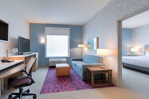 A hotel room with a desk, chair, couch, bedside table, lamp, and a bed. - Home2 Suites By Hilton Alcoa Knoxville Airport (Alcoa)
