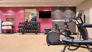 Un gimnasio con equipo de ejercicio, un televisor montado en la pared y un estante con mancuernas.