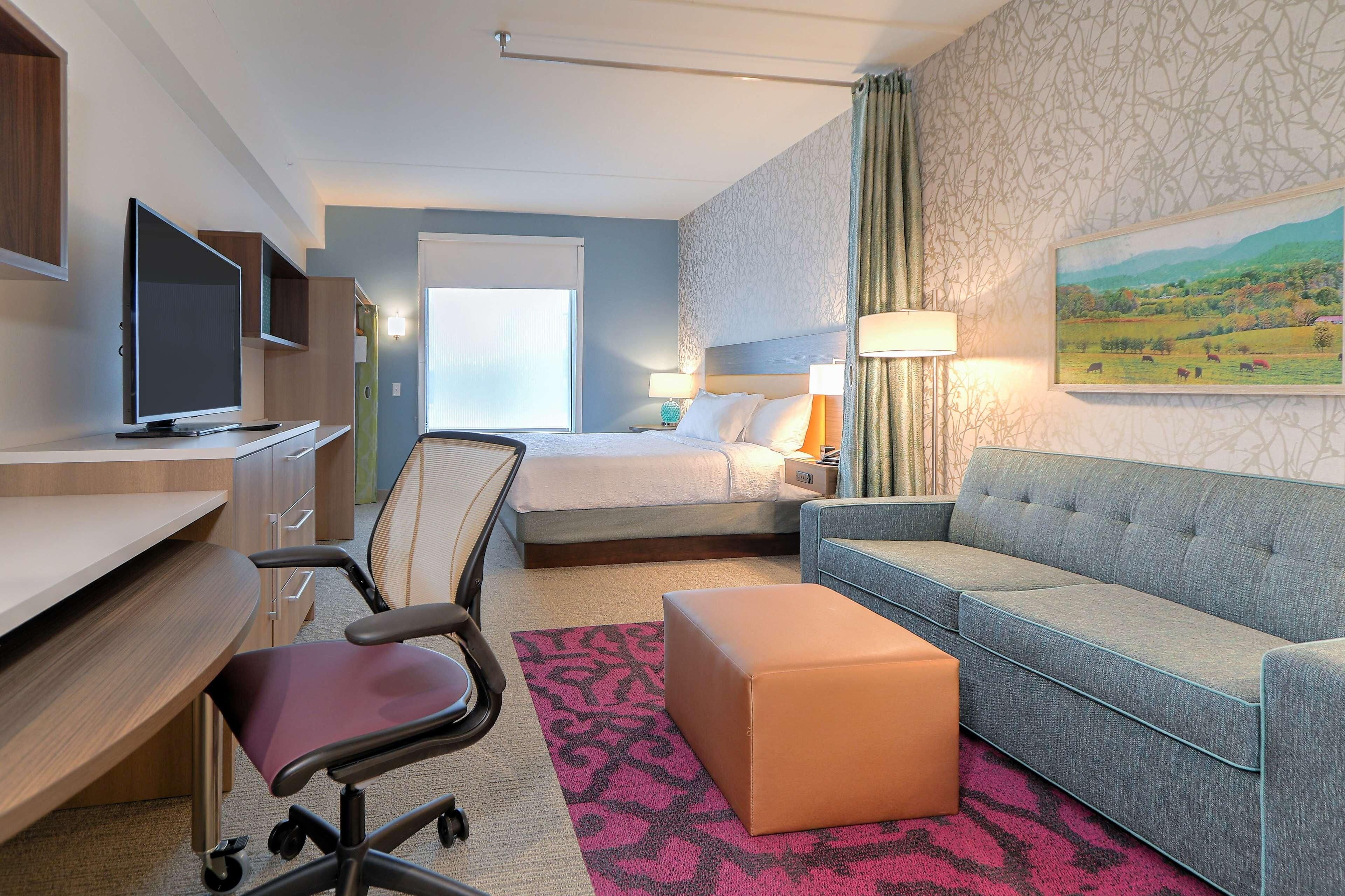 A hotel room with a bed, a sofa, a desk with a computer, a TV, and a painting on the wall.