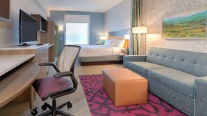 A hotel room with a bed, a sofa, a desk with a computer, a TV, and a painting on the wall. - Home2 Suites By Hilton Alcoa Knoxville Airport (Alcoa)