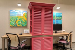 A hotel reception desk with a red cabinet, two office chairs, a computer monitor, and a painting of a field with sunflowers. - Home2 Suites By Hilton Alcoa Knoxville Airport (Alcoa)