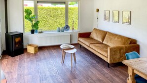 A cozy living room with a sofa, a small table, a wooden floor, a radiator, and a window with a view of a hedge. - Stunning (sta)caravan in Tzummarum (Tzummarum)