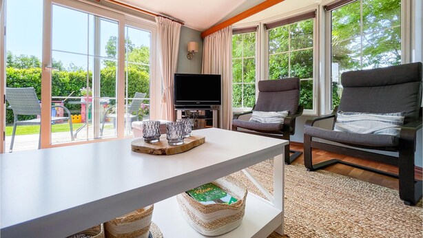 Een moderne woonkamer met een flatscreen televisie, twee grijze fauteuils, een bijzettafeltje met decoratieve objecten en grote ramen met uitzicht op het omliggende landschap.