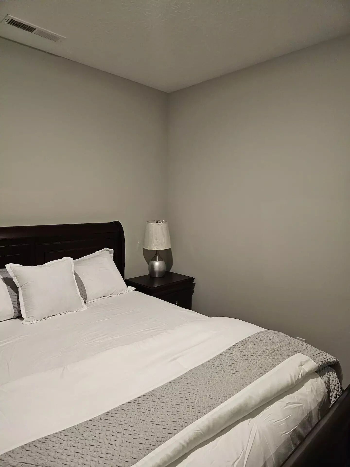 A neatly made bed with white linens and a gray patterned bedspread, a bedside table with a lamp, and a plain wall with a vent.