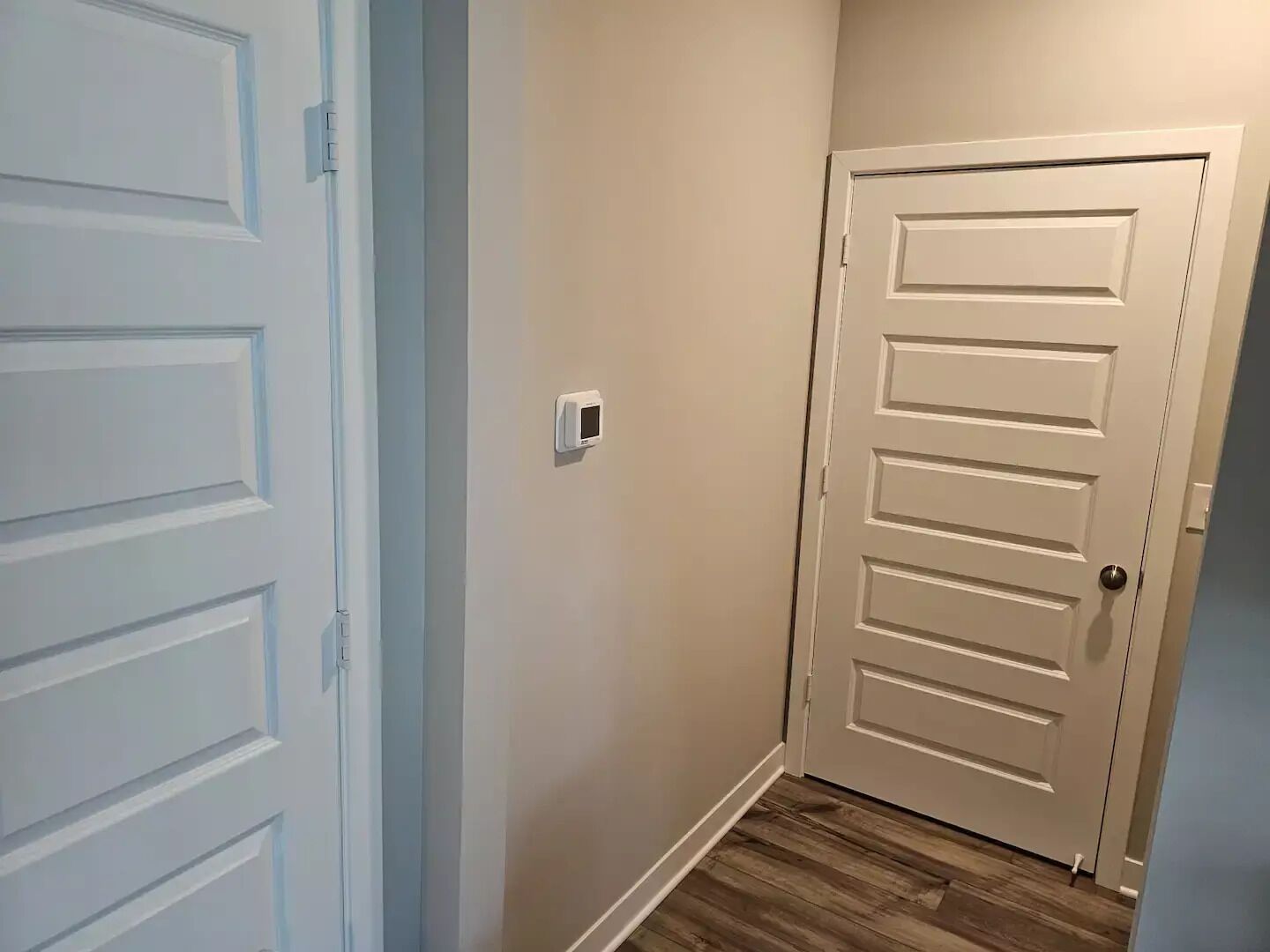 A hallway with a white door, a thermostat on the wall, and wooden flooring.